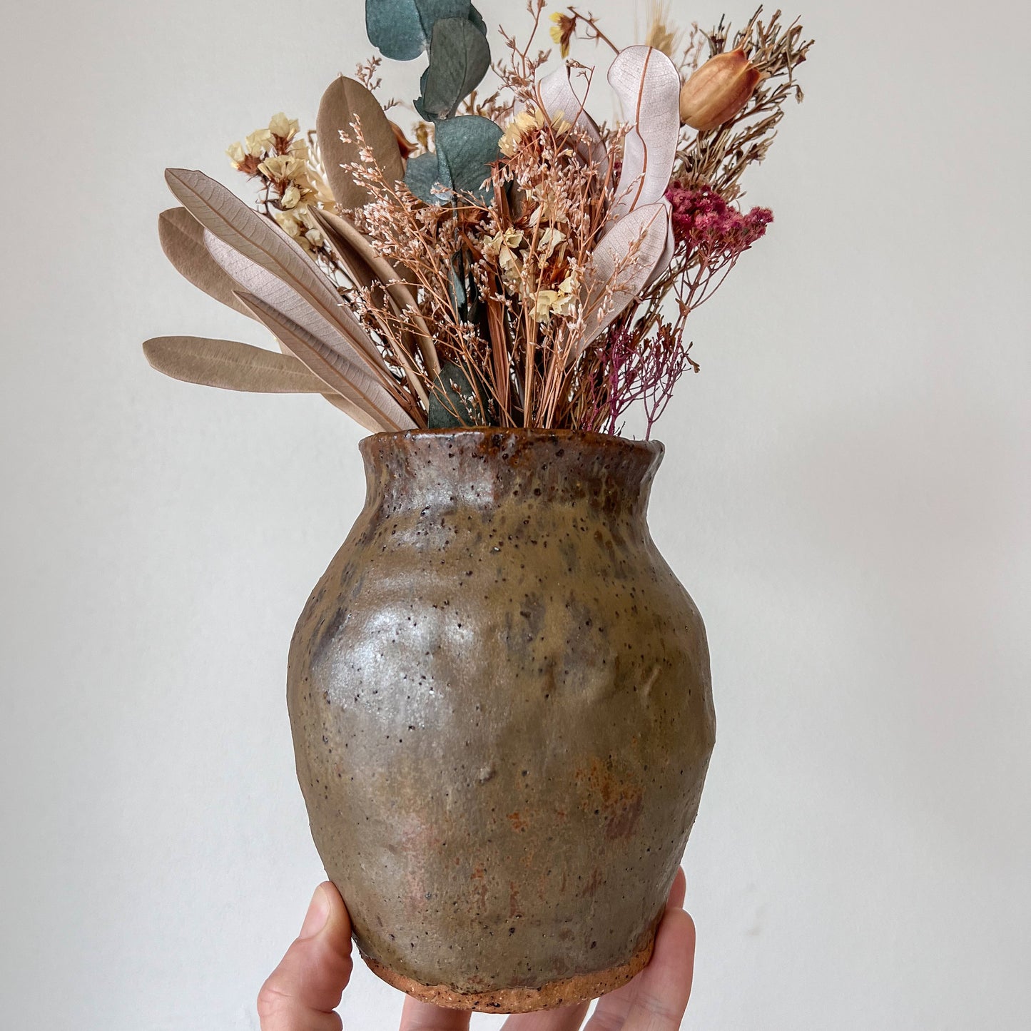 Hand holding a rustic ceramic vase with dried flowers against a plain background
