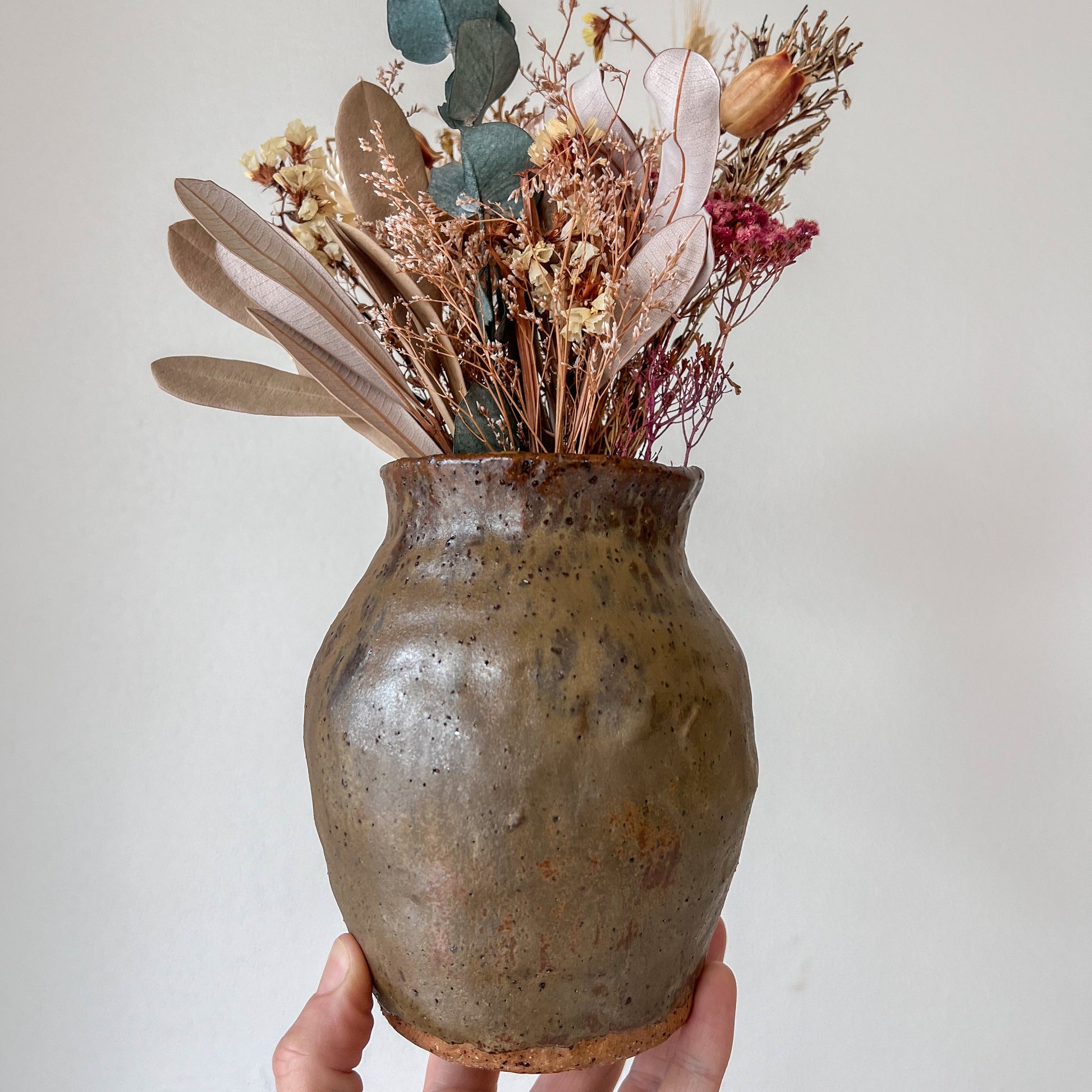 Hand holding a rustic ceramic vase with dried flowers against a plain background