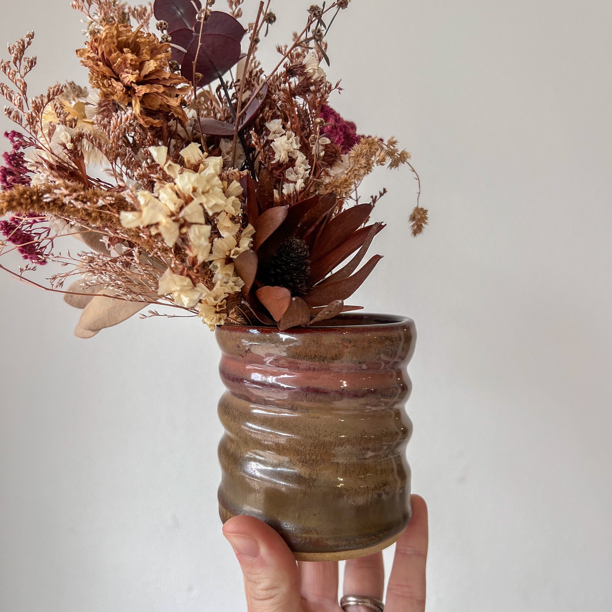 Hand holding a small brown ceramic vase with dried flowers against a plain background