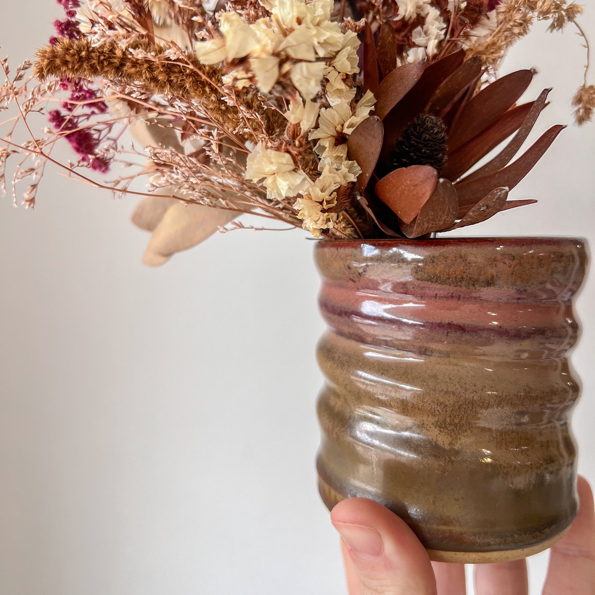 Hand holding a ceramic vase with dried flowers against a neutral background