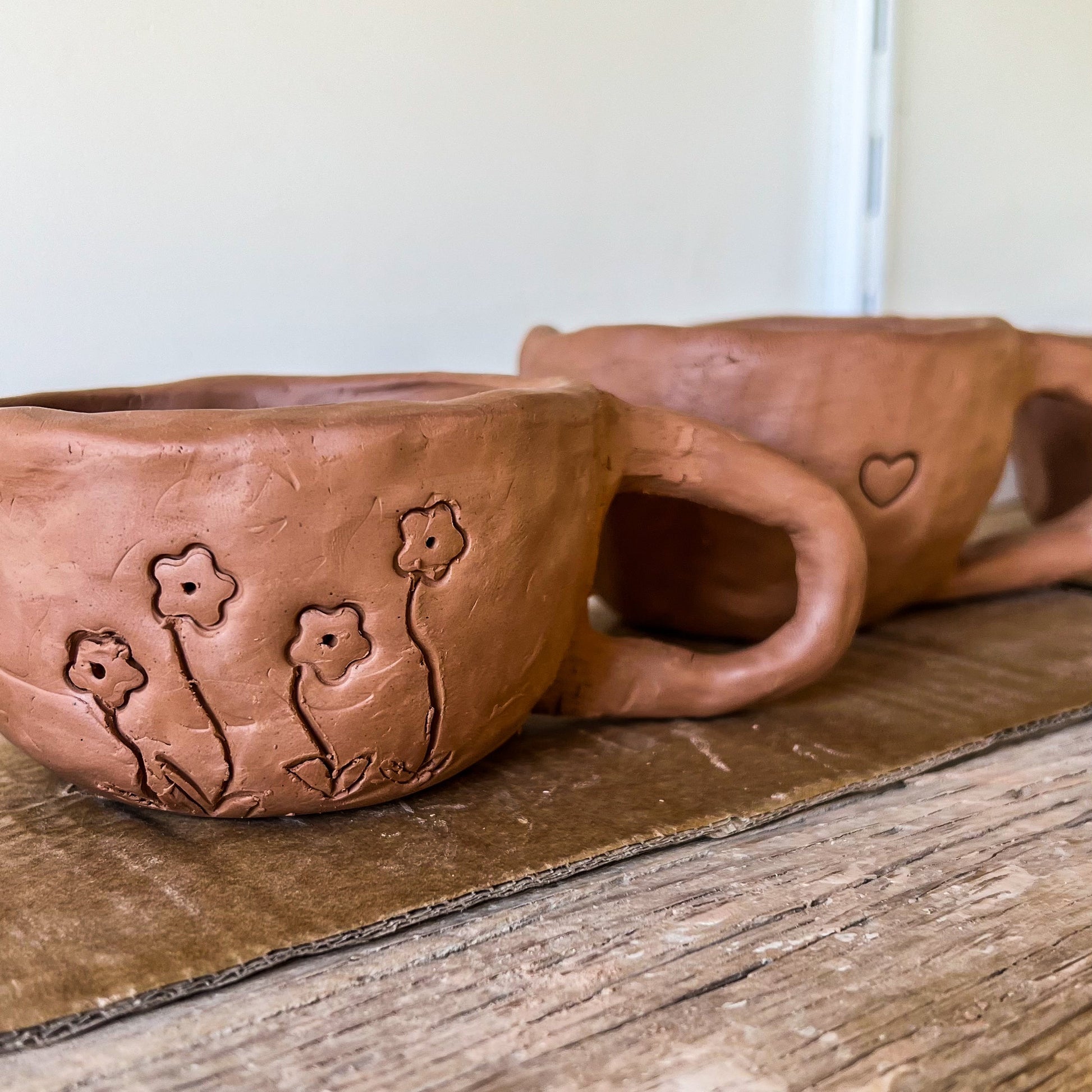 Two clay mugs with floral designs on a wooden surface created at a potter workshop