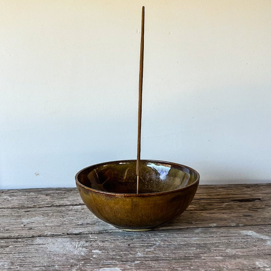 Brown ceramic bowl with a stick inside on a wooden surface against a white wall.