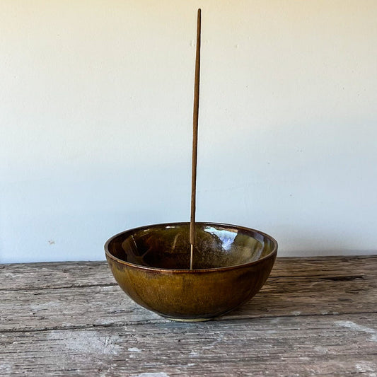 Brown ceramic bowl with a stick inside on a wooden surface against a white wall.