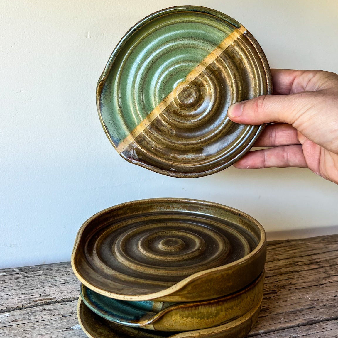 Hand holding a green ceramic plate above a stack of similar plates on a wooden surface.