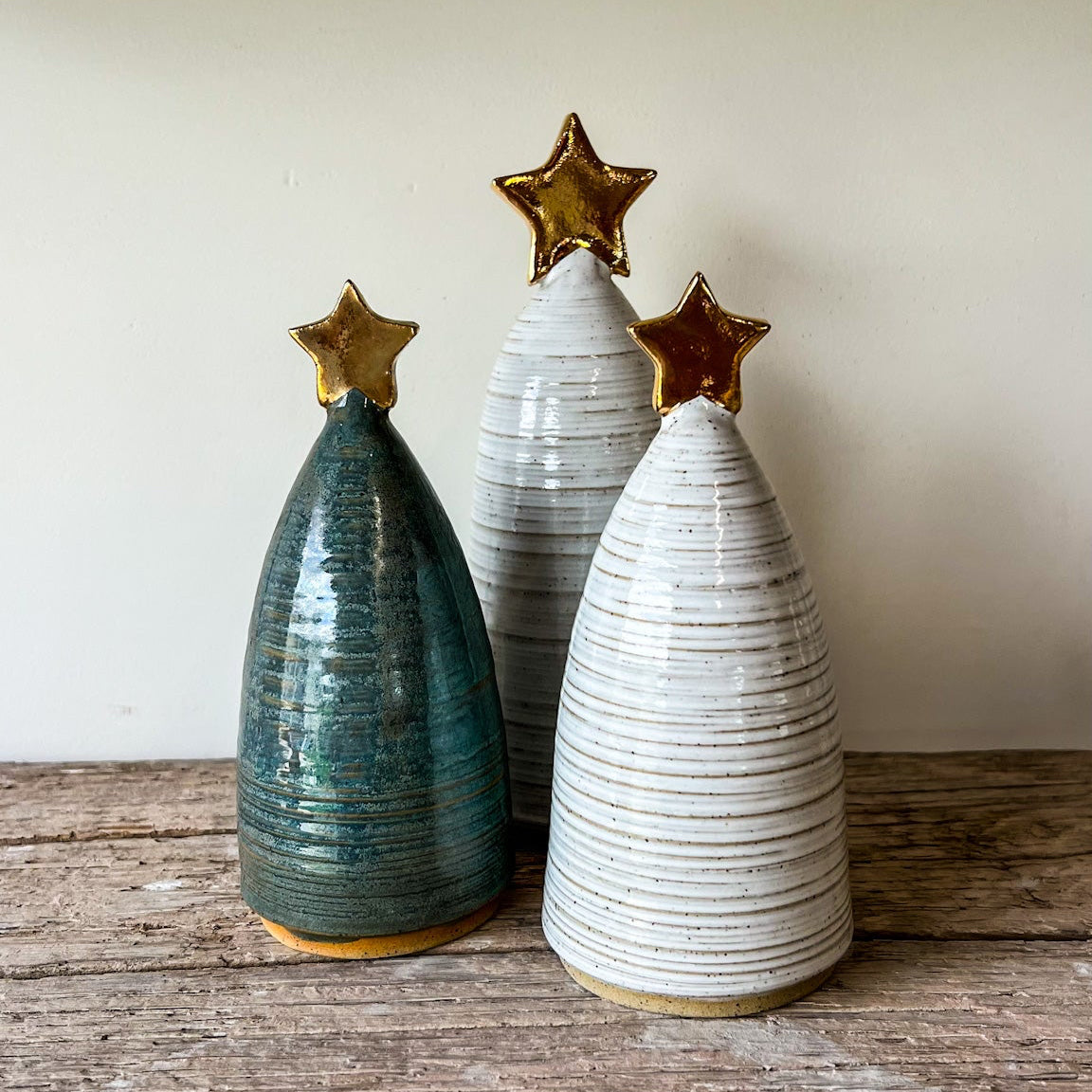 Three ceramic Christmas trees with gold stars on a wooden surface against a beige wall.